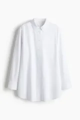 Camisa blanca de algodón con corte holgado, cuello clásico y botones frontales. Presenta mangas largas con puños abotonados y bajo redondeado.