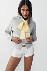 Chaqueta de punto con capucha, manga larga y cierre frontal con cremallera. Presenta un diseño de lazo decorativo en color amarillo en la zona del cuello.
