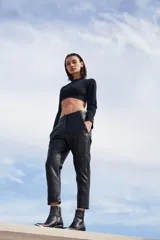 Pantalón sastrero de ecocuero negro, con corte recto, bolsillos delanteros y traseros, y botamanga irregular más corta en la parte frontal.