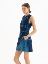 Vestido corto de denim azul con estampa cyanotype de efecto deslavado, sin mangas, escote redondo y cierre frontal completo de metal. Cuenta con corte en la cintura y silueta evasé.