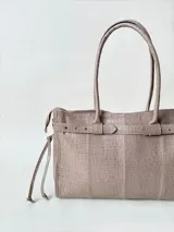 Bolso grande de cuero marrón con textura tipo cocodrilo, estructura rígida y asas para llevar al hombro. Tiene una tira con tachas y hebillas en cada extremo, también lleva lazos en los costados.