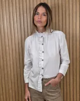 Camisa Magda de jean lino color blanco, con cuello alto y volados sutiles a lo largo de la botonera frontal. Presenta frunces en los hombros y mangas largas.