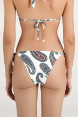 Calzón de bikini con tiras laterales ajustables y diseño estampado con patrón paisley en tonos azules, marrones y naranjas sobre fondo blanco. Cuenta con apliques metálicos decorativos en los cordones.