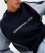 Buzo de corte boxy con cuello redondo y mangas largas raglán. Presenta un diseño bicolor en azul marino y gris, con detalles de piping en contraste y lettering bordado en el pecho.