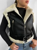 Chaleco negro de cuero sintético con interior de piel sintética color crema, cierre frontal con cremallera, bolsillos laterales con cremallera, cinturón con hebilla metálica y cuello con solapas amplias.