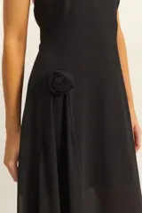 Vestido largo negro con escote halter y espalda descubierta. Presenta un diseño asimétrico con una capa superpuesta de tela semitransparente que cae lateralmente, creando un efecto de movimiento y elegancia.