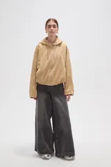 Campera de suede color beige con capucha, cierre frontal completo y mangas largas. Presenta un diseño holgado con bolsillos laterales y detalles de frunces en el ruedo.
