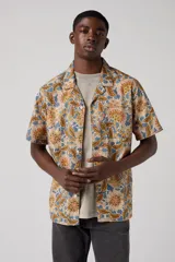 Camisa de manga corta con estampado floral en tonos beige, marrón y azul. Tiene cuello tipo camp y un bolsillo en el pecho.