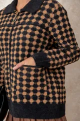 Chaqueta de punto grueso con patrón geométrico de cuadros en blanco y negro. Presenta cuello camisero, cierre frontal con cremallera y tirador circular, bolsillos frontales y puños acanalados.