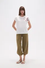 Remera blanca de algodón con mangas cortas y cuello redondo. Presenta un bordado floral en tonos rosados y rojizos en la zona del hombro derecho.