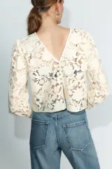 Camisa de encaje color crema con cuello redondo, mangas largas abullonadas y puños ajustados. Presenta cierre en la espalda con botones dorados.