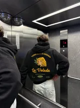 Canguro negro de corte oversize con capucha y bolsillo frontal. Presenta un diseño gráfico estampado en la espalda que simula un paquete de yerba mate, con la ilustración de una mujer, hojas y la inscripción 'La Volada'.