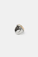 Anillo maxi de metal con engaste ovalado. El engaste tiene un borde exterior trenzado en tono oscuro y un borde interior liso en tono dorado, sosteniendo una piedra grande facetada de color transparente.