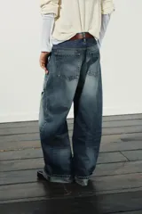 Jeans de mezclilla azul con lavado desgastado y rotos en la pierna derecha. Tienen tiro medio, corte ancho (baggy) y puños doblados en el bajo.