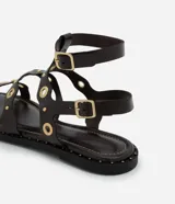 Sandalia plana tipo gladiadora color negro, confeccionada en poliuretano. Presenta tiras onduladas con recortes ahuecados y detalles de ojalillos y tachas metálicas doradas. Cuenta con doble tira ajustable en el tobillo con hebillas metálicas.