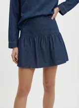 Pollera corta de denim azul medio, con tiro medio y cierre de cremallera. Presenta un diseño con un volante fruncido en el ruedo.