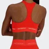 Top deportivo de soporte medio, color naranja brillante, con escote redondo y espalda tipo nadador. Presenta una banda elástica ancha bajo el busto con el logo de New Balance repetido en blanco.