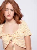 Top corto de manga corta, color blanco con estampado de cuadros pequeños en naranja. Presenta escote redondo con fruncido y lazo ajustable en el frente.