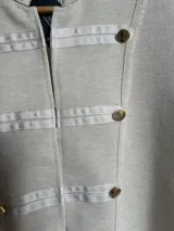 Chaqueta de algodón color beige de estilo militar, con cuello mao, detalles de bandas horizontales en tono más claro y botones dorados decorativos en el frente y puños.