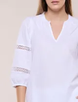 Blusa blanca de calce suelto y caída ligera, con escote en V y cuello Mao pequeño. Presenta mangas tres cuartos abullonadas con elástico en los puños y detalles de calado o puntilla encastrada. Espalda lisa con canesú y leve frunce.