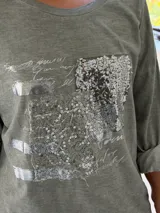 Remera de manga larga color verde, con cuello redondo y diseño holgado. Presenta un detalle frontal con parches de lentejuelas plateadas y estampado tipográfico.