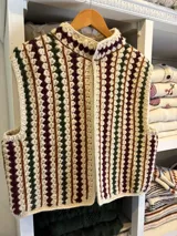 Chaleco tejido a mano en crochet de lana merino color negro, con diseño sin mangas, cuello mao y cierre delantero con cordones para atar. Presenta un patrón de rayas verticales con detalles geométricos en tonos fucsia, celeste y blanco.