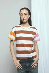 Remera de manga corta con diseño a rayas horizontales en blanco y marrón, con detalles de costuras decorativas en zig-zag de color verde. Las mangas presentan bloques de color naranja en un lado y rosado en el otro.
