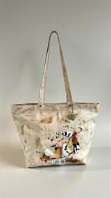 Cartera tipo tote bag de cuero natural con diseño artístico pintado a mano, doble asa de hombro y detalle de dije colgante metálico.