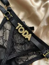 Tanga de tul translúcido negro con diseño de encaje tipo monograma, tiras elásticas regulables con argollas doradas y detalle de letras en strass formando la palabra "TODA".