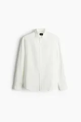 Camisa blanca de corte regular fit, confeccionada en una mezcla suave de lyocell y algodón. Presenta cuello inglés, tapeta clásica, bolsillo superior abierto en el pecho y canesú con pliegue de caja en la espalda. Mangas largas con abertura y puños ajustables con botón. El bajo es redondeado.