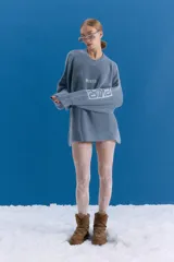 Sweater de punto en color azul grisáceo con diseño oversize. Presenta cuello redondo, mangas largas y detalles de estampados gráficos en el frente y en una de las mangas. El tejido posee un acabado desgastado que le otorga un aspecto vintage.