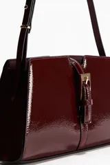 Cartera bandolera color bordó de cuero charolado, con correa fina ajustable y cierre metálico dorado en la parte frontal. Cuenta con un bolsillo interior con cierre y está forrada.