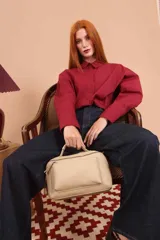 Bandolera de diseño rectangular en color suela, confeccionada en material sintético. Cuenta con un asa superior fija, correa larga ajustable y extraíble, y dos bolsillos frontales con cierre metálico, uno de ellos con diseño superpuesto. Presenta el logo de la marca grabado en el frente.