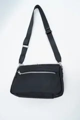 Bandolera de diseño compacto en color negro, confeccionada en material sintético resistente. Cuenta con un compartimento principal con cierre y un bolsillo frontal con cierre metálico visible. Incluye una correa larga ajustable y extraíble para llevar al hombro o cruzada.