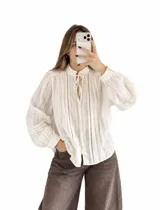 Blusa blanca de estilo romántico, confeccionada en tela liviana con detalles de alforzas y encaje insertion en el frente. Cuenta con cuello alto fruncido con lazo de ajuste, mangas largas abullonadas con puños elásticos y cierre frontal con botones.
