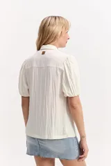 Camisa de manga corta color blanco marfil con textura y mangas abullonadas. Tiene cuello camisero, calce suelto y cierre frontal con botones.