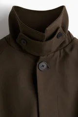 Gabardina estilo Mac de color marrón oscuro, confeccionada en sarga suave. Presenta cuello clásico, cierre frontal con botones ocultos, manga raglán con trabilla y botón en los puños, bolsillos insertados y cinturón extraíble para atar a la cintura. Tiene una abertura en la espalda y forro interior.