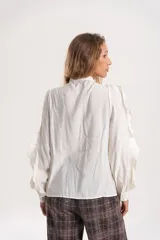 Camisa de gasa color blanco con volados frontales y en las mangas, cuello con lazo y mangas largas abullonadas con puños ajustados.