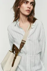 Bolso tipo saca color crudo con cuerpo en tejido canvas, asa corta de mano trenzada en marrón y correa bandolera adicional. Cuenta con cierre de botón imantado y bolsillo interior con cremallera.