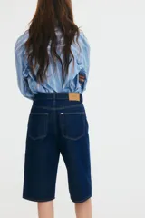 Bermuda de denim azul oscuro, tiro alto, corte ancho, largo a la rodilla y cinco bolsillos. Cierre frontal con botón.