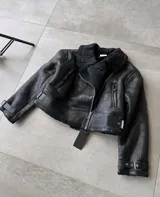 Campera tipo gamulán de cuero sintético negro con interior y cuello de corderito sintético. Presenta cierre frontal, bolsillos laterales con cierre y correas ajustables con hebillas en los puños y en la cintura.