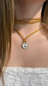 Gargantilla tipo choker compuesta por una tira de gamuza color mostaza que rodea el cuello y un dije metálico en forma de candado con diseño de corazón calado.