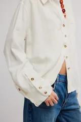 Blusa de lino color blanco crudo, de corte relajado, con cuello clásico, cierre frontal de botones, bolsillo en el pecho y mangas largas con presillas para arremangar.