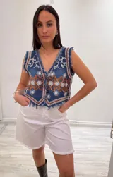 Chaleco corto en denim azul con detalle desflecado y bordado artesanal en tonos celeste y dorado. Estilo boho chic con escote en V, ideal para combinar con prendas básicas y elevar tu look.