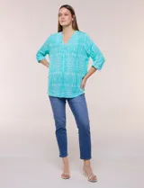 Blusa turquesa de silueta suelta confeccionada en tejido liviano, con escote en V, pequeña cartera frontal con botones decorativos y mangas 3/4 con presilla para arremangar. Presenta patrón estampado geométrico tipo tie-dye y ruedo curvo.
