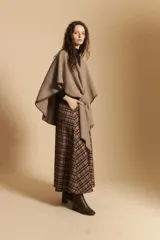 Poncho de tejido suave color beige con diseño envolvente y terminaciones con costuras decorativas en contraste color negro. Presenta un detalle de lazo frontal integrado.
