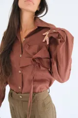 Camisa blanca de manga larga, con botones de guampa y un detalle de roseta o flor a tono en la parte delantera. El calce es holgado con la espalda ligeramente más larga.