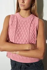 Chaleco sin mangas de tejido de punto color rosado, con cuello redondo y punto trenzado (ochos) en el frente. Presenta un borde acanalado en el ruedo.