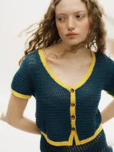 Top de tejido calado con diseño de punto abierto, color azul petróleo con detalles en amarillo en cuello, puños y ruedo. Presenta escote en V, cierre frontal con botones tipo carey y mangas cortas.