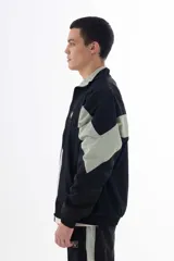 Campera negra de nylon con recortes en forma de V en color gris en las mangas. Tiene cuello mao, cierre frontal, puños y cintura elásticos, bolsillos laterales y logo estampado en el pecho y en la manga izquierda.